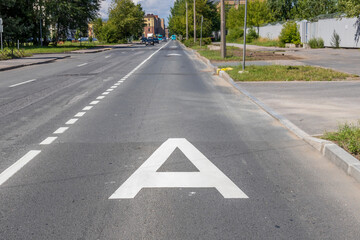 A white letter A painted on the asphalt of a city street, marking a designated lane. The street is surrounded by sidewalks and a grassy area. The road is empty except for the white markings..