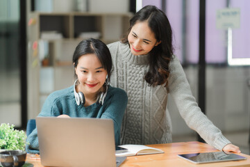 Collaborative Learning: Two young Asian women engage in focused teamwork, utilizing a laptop for a shared project in a modern office setting. 