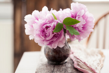 Beautiful freshly cut pink peonies in a vintage copper vase on old wooden board, linen runner and a vintage Windsor chair in the background © Syphoto