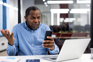 Confused office worker holding phone with puzzled expression at desk with laptop and tablet. Image...