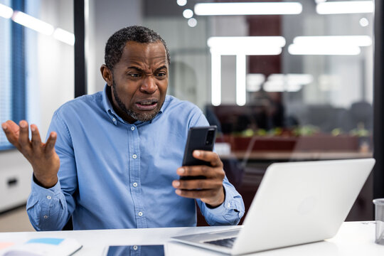 Confused office worker holding phone with puzzled expression at desk with laptop and tablet. Image depicts frustration, confusion, and technology issues in modern business environment. - Powered by Adobe