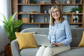 Smiling senior woman working on laptop sitting on sofa. Comfortable home setting with bookshelves and plants in background. Concept of remote work, technology, senior lifestyle, happiness.