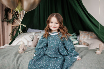 Cheerful little birthday girl 4-5 years in stylish rustic style dress sits on bed decorated with baldachin, pillows and balloons, smiles and looks at camera. Child birthday party celebration. © Andriy Medvediuk