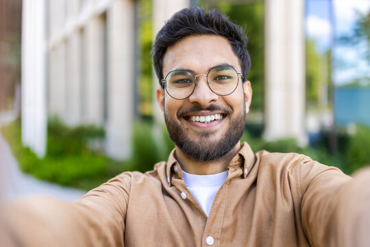 Close-up of smiling man taking selfie outdoors on sunny day. Man with beard and glasses in casual clothing enjoying outdoor moment. Concept of happiness, modern communication, and casual lifestyle.