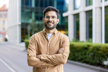 Young man with glasses standing confidently outside modern office building. Smiling and crossing arms, conveying confidence and friendliness. Background features urban environment