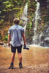 Obraz premium Back, hiking and photography with man at waterfall in nature for adventure, exploration or journey. Environment, jungle and travel with photographer outdoor in forest for hobby, leisure or memory