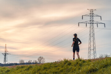 Runner on grassy hill near power lines at sunset. Silhouette of woman jogging against colorful sky. Outdoor exercise and energy infrastructure juxtaposed in scenic landscape