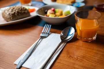 Close-up of cutlery, napkin and orange juice in bright warm light on a nicely set table with bread and fruit salad on the background 