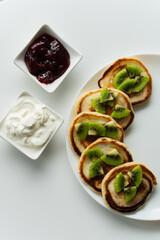 Plate of pancakes dripping with cherry jam, sour cream and kiwi pieces with white background.. Copy space for text. Mockup for menu, Pancake day