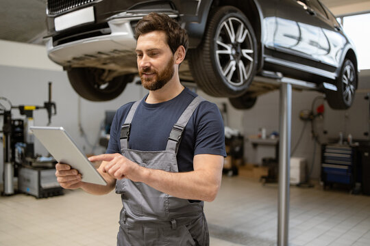 Mechanic using tablet in car repair shop under lifted vehicle. Focused automotive technician inspecting diagnostics, modern technology in auto maintenance, professional service environment.