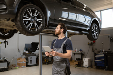 Mechanic in uniform using tablet while inspecting car on lift in garage. Professional workshop...