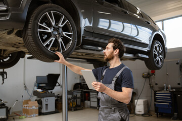 Mechanic using tablet while inspecting car on lift in workshop. Professional car maintenance,...
