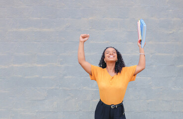 Black Brazilian university student celebrating her success in studies or graduation. They are happy with their arm raised while holding notebooks and folders. In the background we have a wall brick.
