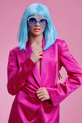 A woman in a vibrant pink suit, wearing blue sunglasses, strikes a confident pose against a pink background with her hand on her hip and eyes closed