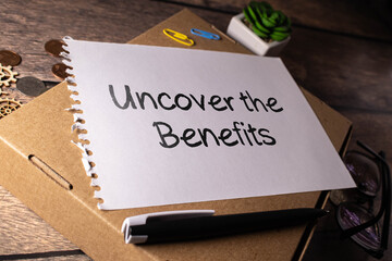 Uncover the Benefits - text written on notebook on wooden table with pen coffee and glasses.