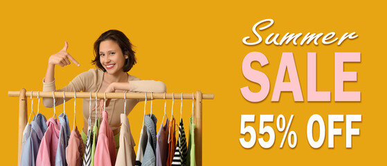 Beautiful young woman pointing at rack with different clothes on yellow background