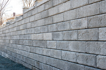 A large grey precast concrete block wall. The massive rectangle shaped bricks are interlocking cinder bricks with granular backfill. The textured castle style wall has large square caps. 