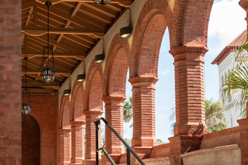 The exterior entrance and hallway to a historical red brick building with columns, high ceiling, an arched corridor, wooden roof and metal light fixtures.The arches are open to the outside of the hall