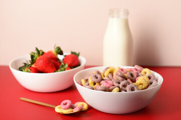 Bowls with colorful sweet cereal rings, strawberries and bottle of milk on red table