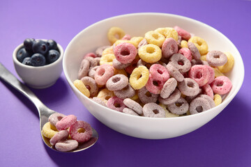 Bowls with colorful sweet cereal rings and blueberries on purple background