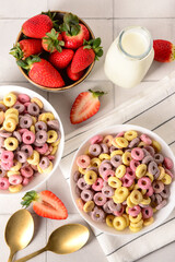 Bowls with colorful sweet cereal rings, strawberries and bottle of milk on white tile table