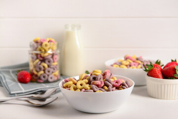 Bowls with colorful sweet cereal rings, strawberries and bottle of milk on white table