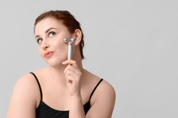 Thoughtful young woman using facial massage tool on grey background