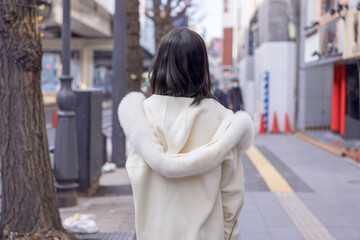 Fototapeta premium Twenty-something Chinese woman with black hair wearing a white coat with a fluffy fur hood and a white skirt walking on a public road in Minato, Tokyo, Japan.