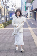 Twenty-something Chinese woman with black hair wearing a white coat with a fluffy fur hood and a white skirt walking on a public road in Minato, Tokyo, Japan.