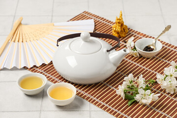 Teapot with cups, spoon of dry tea and flowers on white tile background