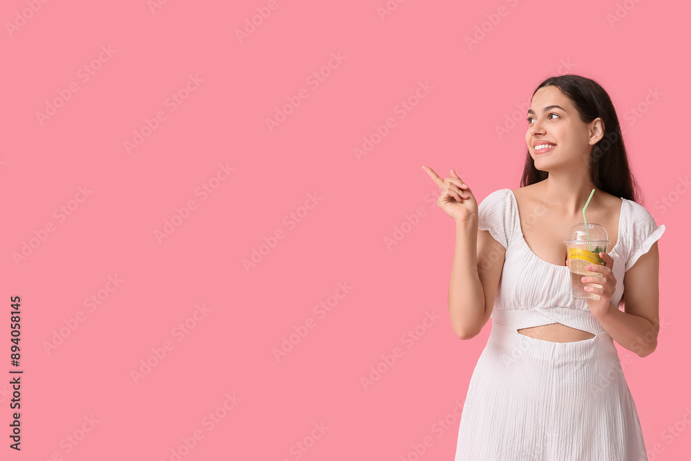 Poster beautiful young happy woman with cup of fresh lemonade pointing at something on pink background