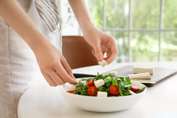 Young woman putting pieces of feta cheese in salad at home