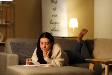 Young woman reading book and resting on cozy sofa in living room at night