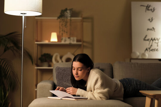 Young woman reading book and resting on cozy sofa in living room at night