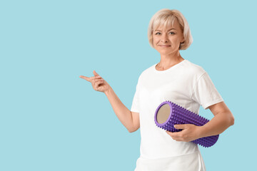 Sporty beautiful mature happy woman with foam roller pointing at something on blue background