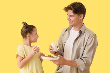 Little girl and her father holding glasses of milk with cookies on yellow background