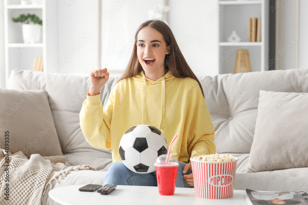 Wall mural beautiful young happy woman with soccer ball, cup of cola and popcorn watching football on tv at hom