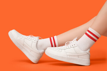 Legs of young woman in stylish white sneakers on orange background