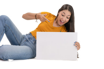 young Latina girl in casual clothes, lying on the floor with a big smile on her face, pointing to blank white poster in front of her.