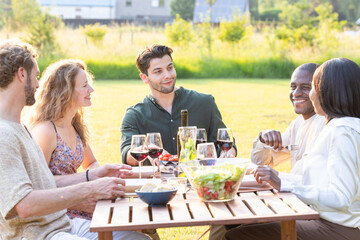 A group of friends is enjoying a meal and wine outside, fostering a relaxed atmosphere in a sunny garden setting