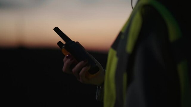 A person holding a walkie-talkie at dusk in a reflective vest