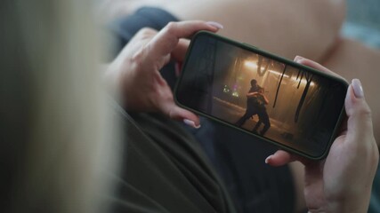 Close up of woman holding mobile phone in hands and watching action movie lying on sofa. Smartphone screen with displayed thriller film. Caucasian woman rests at home on the weekend. Lifestyle concept