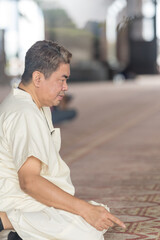 Fifty-something Muslim Malay person in religious attire, wearing a white outfit and a black hat, praying devoutly on a majestic carpet inside a very large mosque in Putrajaya, Malaysia.