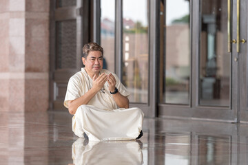 Fototapeta premium Fifty-something Muslim Malay person in religious attire, wearing a white outfit and black hat, praying devoutly in a wide glass-enclosed marble area outside very large mosque in Putrajaya, Malaysia.
