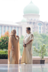 50-something Muslim Malay man in white outfit and black hat and 30-something Chinese woman in cream-colored religious attire standing together outside large mosque in Putrajaya, Malaysia.