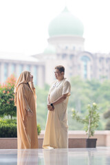 50-something Muslim Malay man in white outfit and black hat and 30-something Chinese woman in cream-colored religious attire standing together outside large mosque in Putrajaya, Malaysia.