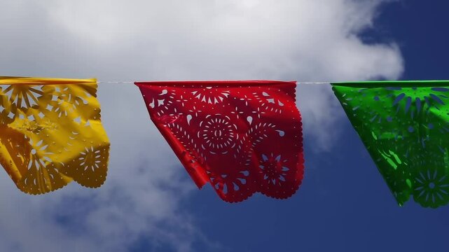 Colorful papel picado banners in yellow, red, and green against a backdrop of clouds and blue sky. These intricately cut paper decorations are traditionally used in Mexican festivities.