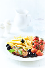 Roast Pork Steak with french fries, tomatoes and olives. White wooden background. Close up.	