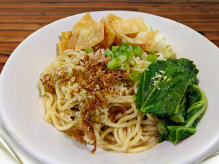 Close up shot of Indonesian chicken noodles on a white bowl. It has shredded chicken, fried skin dumplings, onion, fried shallots, and choy sum as toppings.