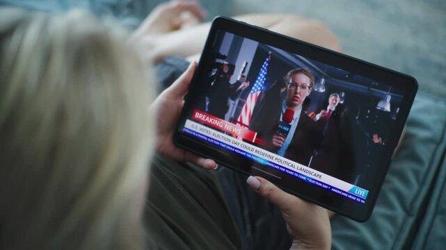 Close up of Caucasian woman watching breaking news broadcast about US Election Day using tablet computer while on sofa. Playback of news report with female journalist displayed on the tablet screen.